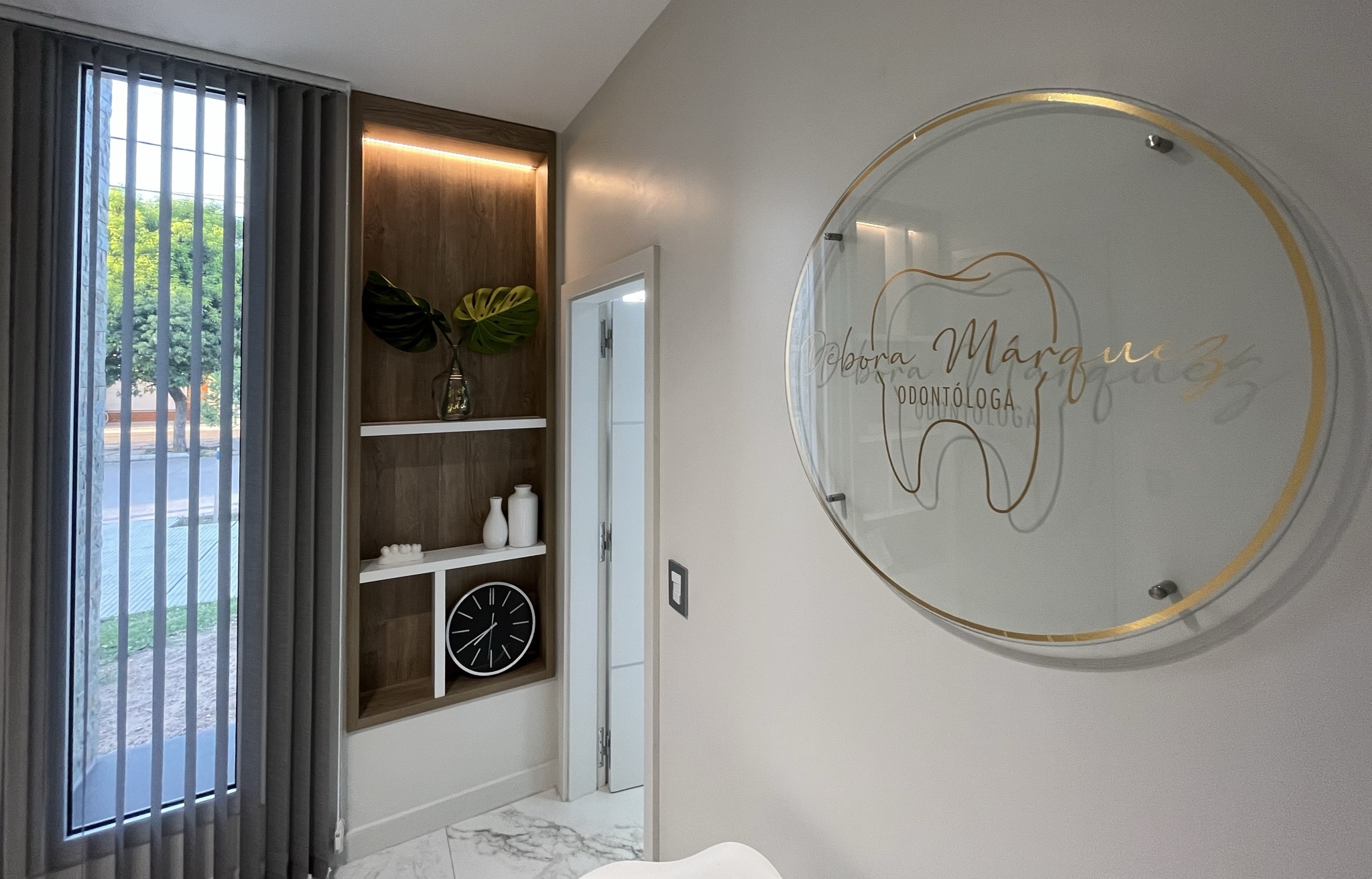 foto de la sala de espera de pacientes del consultorio
                de Débora Márquez, en la misma se observan sillones, 
                cartel con de vidrio con logo, ventana con cortinas 
                y repisas decorativas con luces