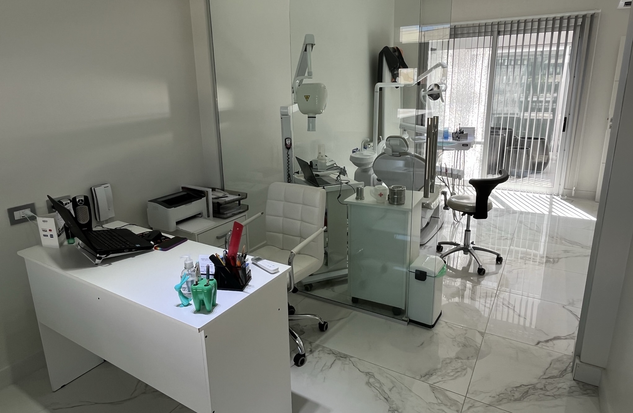 foto de la oficina de la Débora Márquez, se observa su 
                            escritorio color blanco, junto a su compuradora y utiles laborales, 
                            al fondo se ve el sillon odontológico junto a su equipamiento