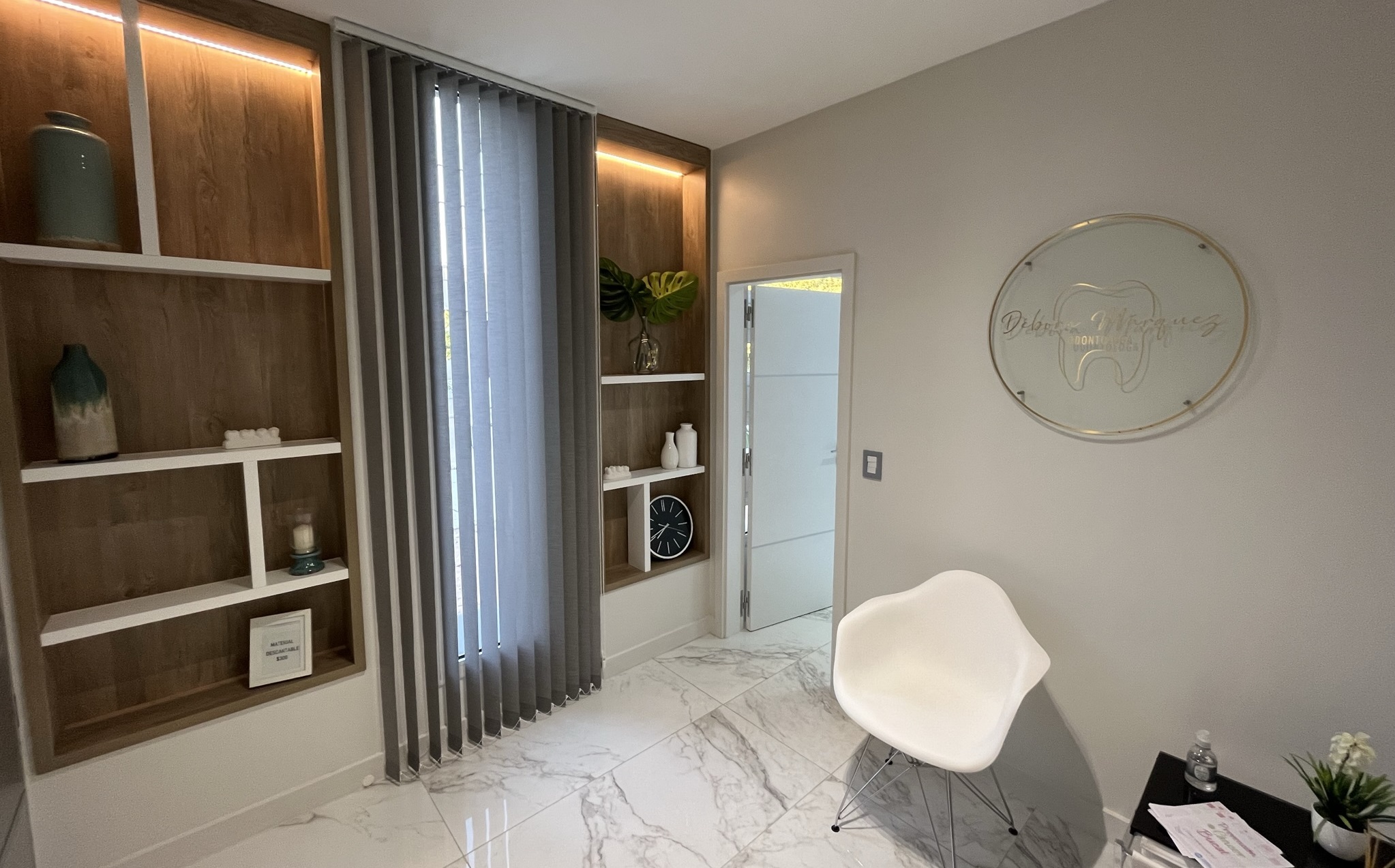 foto de la sala de espera de pacientes del consultorio
                            de Débora Márquez, en la misma se observan sillones, 
                            cartel con de vidrio con logo, ventana con cortinas 
                            y repisas decorativas con luces